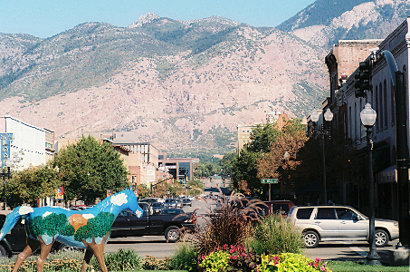 [A horse statue with colored scenery painted on it sits amid some manicured plants. This view looks down a wide street with two to three story buildings on either side. The mountains at the back end of the town are brown with sections with green trees. The mountains are huge and completely dwarf the buildings.]